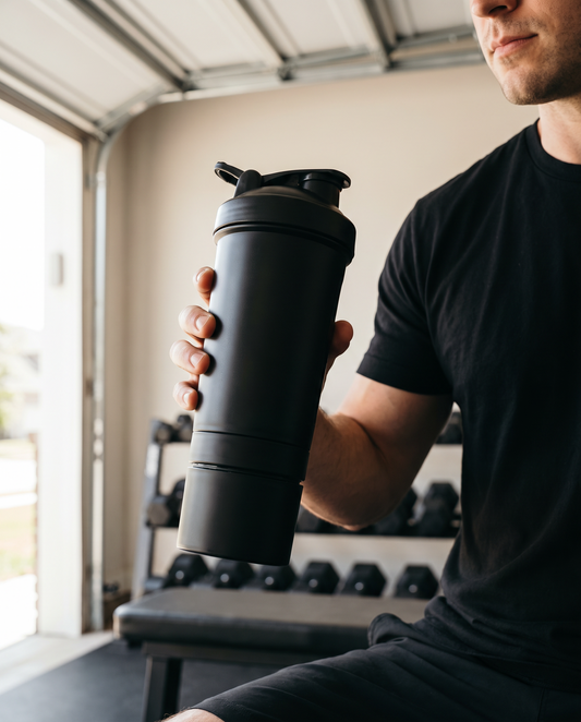 Black three-layer protein shaker bottle with twist-off storage base held in a garage gym